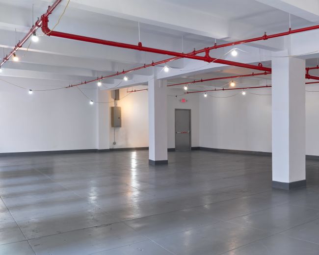 The image shows a spacious, empty room with polished gray flooring, white walls, and exposed red piping along the ceiling.