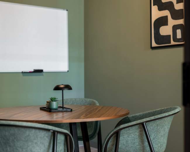 A small meeting room features a round wooden table surrounded by three green chairs, with a whiteboard on the wall and an abstract painting nearby.