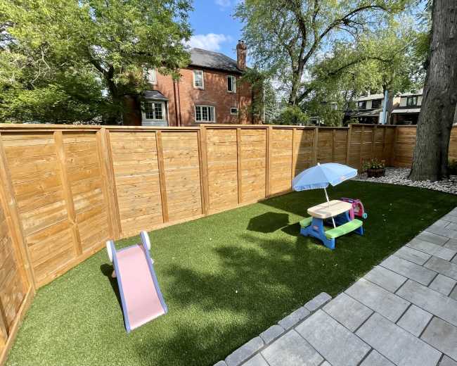 A small, fenced backyard features artificial grass, a slide, a play table, a pink backpack, and a blue umbrella.