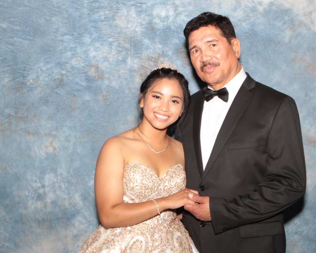 A young woman in a formal gown poses with a man in a tuxedo against a textured blue background.