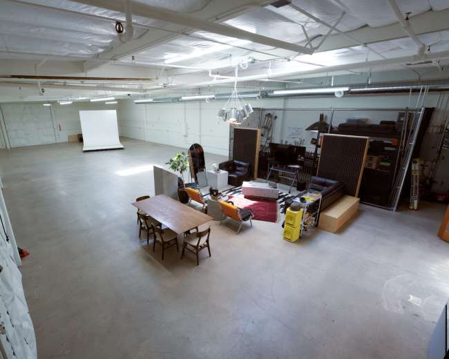 The image shows a large, empty industrial space with concrete floors, a white projection screen at the far end, and various furniture pieces scattered around, including a table and chairs.