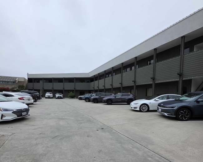 The image shows a parking lot with several cars arranged along both sides of a concrete lane beside a two-story building.