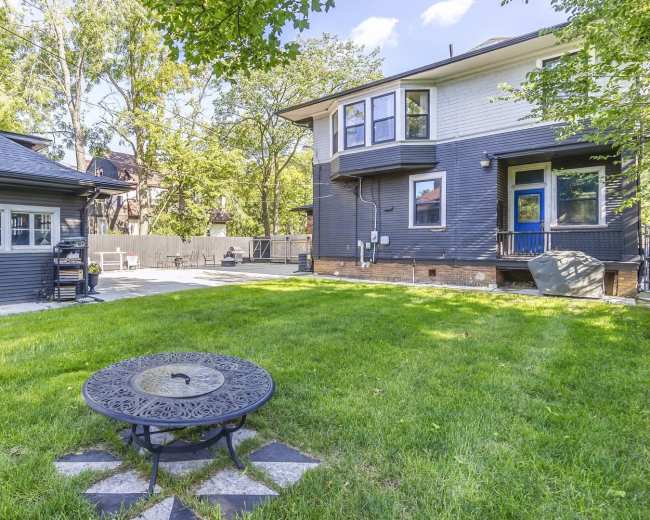 The image shows a backyard featuring a circular metal table on a stone patio, surrounded by a green lawn and a two-story house with a deck and a separate garage.