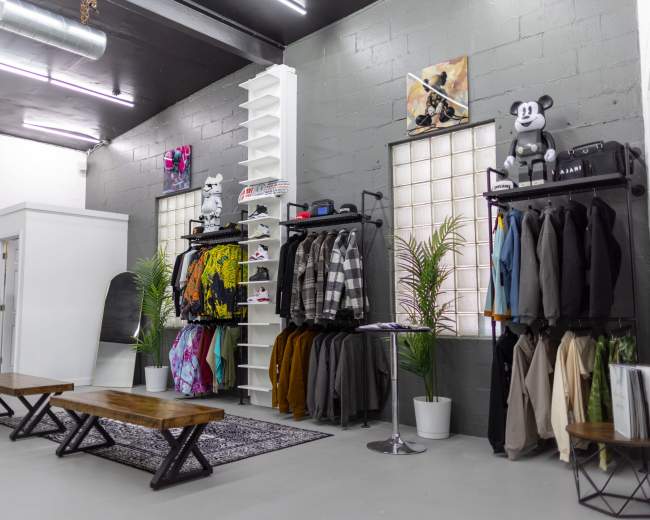 The image shows a modern retail space featuring clothing racks with various garments, benches for seating, and decorative plants against gray walls.