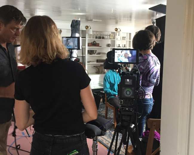 A film crew sets up equipment in a brightly lit room with a table, preparing for an interview scene.