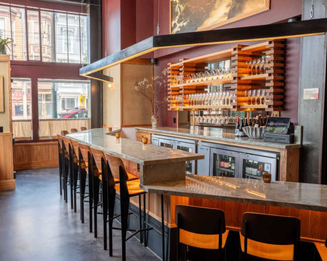 The image features a modern bar area with a sleek marble countertop, wooden shelves holding bottles, and seating along the side facing large windows.