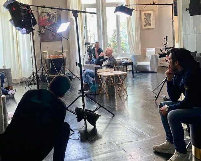 A group of people is sitting in a well-lit room with film equipment set up for a shoot.