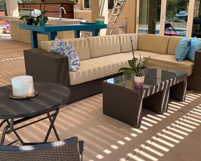 A modern outdoor seating area features a beige sofa set with colorful cushions and a glass coffee table on a patio with shaded patterns from nearby structures.