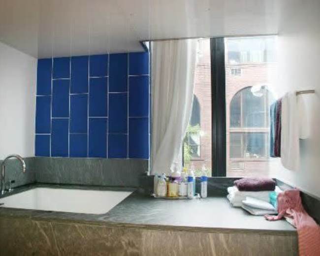 A modern bathroom with a large soaking tub, a marble countertop, and a blue tiled wall, featuring a window with sheer curtains.