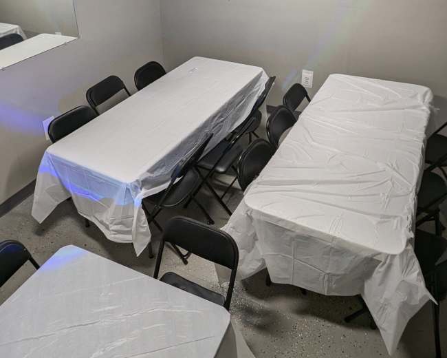 The image shows a room with several rectangular tables covered in white tablecloths, surrounded by black chairs.