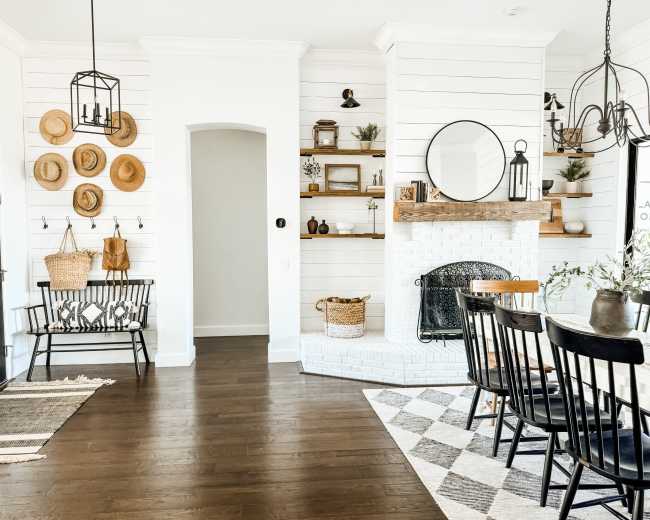 The image shows a spacious, well-lit room featuring a dining area with a patterned rug, a black table surrounded by chairs, and a cozy fireplace with decor and seating elements nearby.