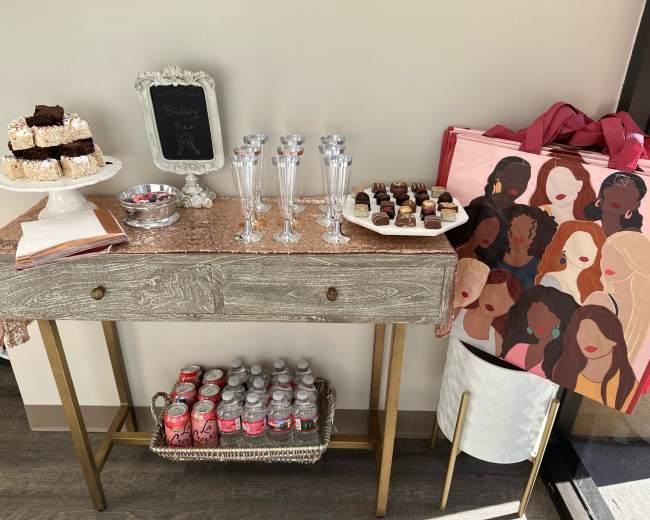 A table decorated with a pink cloth features desserts, sparkling glasses, and a decorative bag, set against a wall.