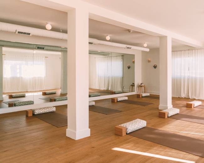 The image shows a bright yoga studio with wooden floors, several yoga mats laid out, and large mirrors reflecting the space.