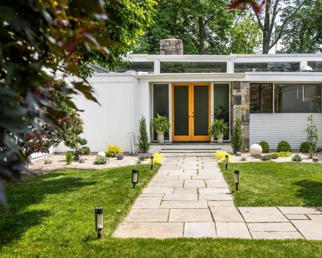 The image shows a modern home entrance with double yellow doors, surrounded by a landscaped garden featuring stone pathways, plants, and lighting fixtures.