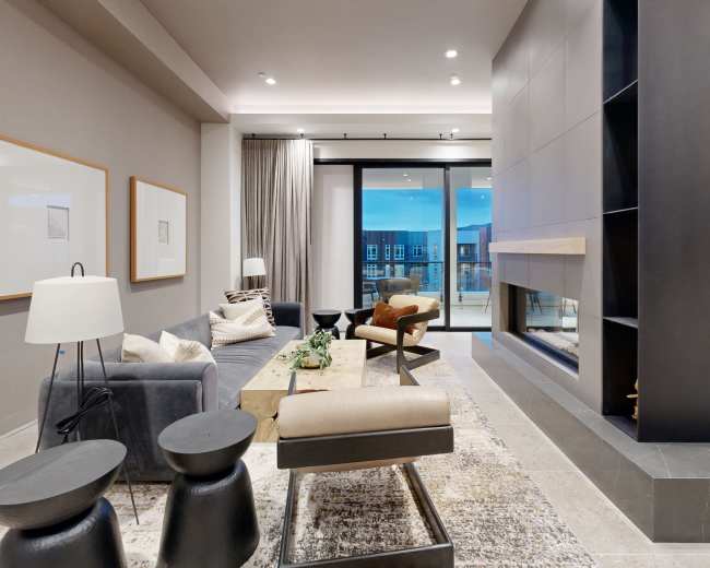 The image shows a modern living room featuring a sectional sofa, a coffee table, a fireplace, and large glass doors leading to a balcony.