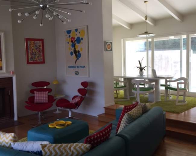 The image shows a modern living room with a turquoise sofa, red chairs, and a dining area featuring a table and chairs, accented by large windows and colorful artwork.