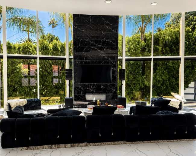 The image shows a modern living room with a large black sectional sofa, a sleek marble floor, and a wall-mounted television surrounded by large windows offering a view of lush greenery.