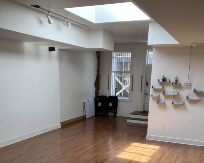 A bright, empty room with hardwood floors features a skylight and a small entry area with two chairs and wall-mounted shelves.