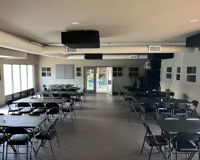The image shows a spacious room set up with black folding tables and chairs arranged in a grid layout, with large windows on one side and double doors leading to another space.