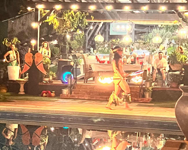 A woman twirls flaming poi beside a pool reflecting lights and a decorated outdoor space filled with people and greenery.