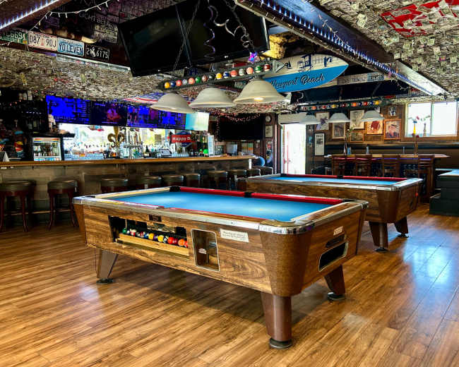 The image shows a bar area with two pool tables, wooden flooring, and a ceiling decorated with various items, including photographs and license plates.