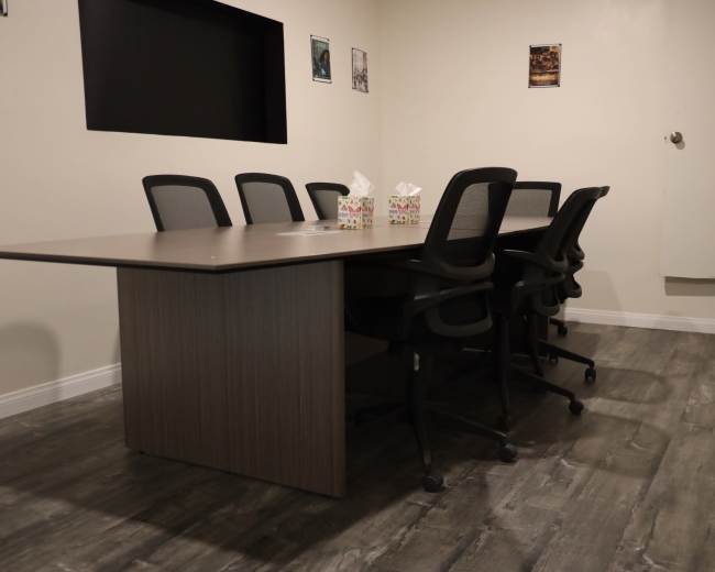 The image shows a conference room with a large table surrounded by several ergonomic chairs and a wall featuring a black screen and framed pictures.