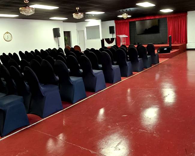 The image shows a spacious indoor auditorium with rows of black chairs facing a stage, which is illuminated by overhead lights and features red curtains and a large screen.