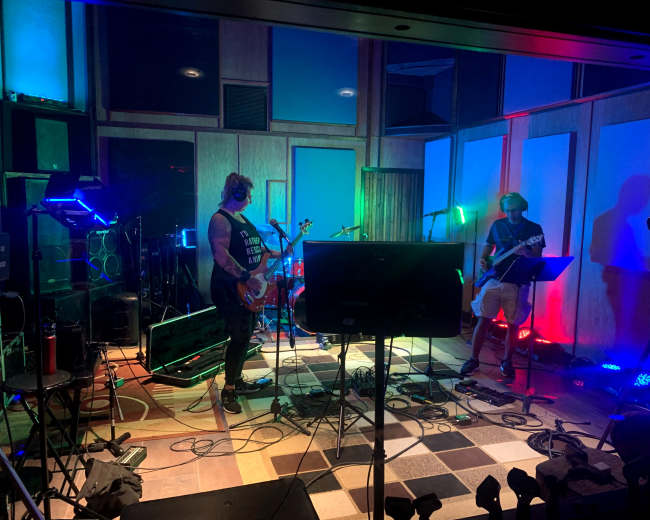 Two musicians perform in a recording studio equipped with colorful lighting and various instruments.