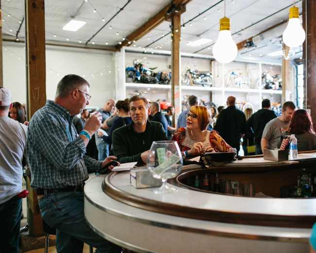 A group of people converse at a circular bar in a busy venue filled with motorcycles on display.
