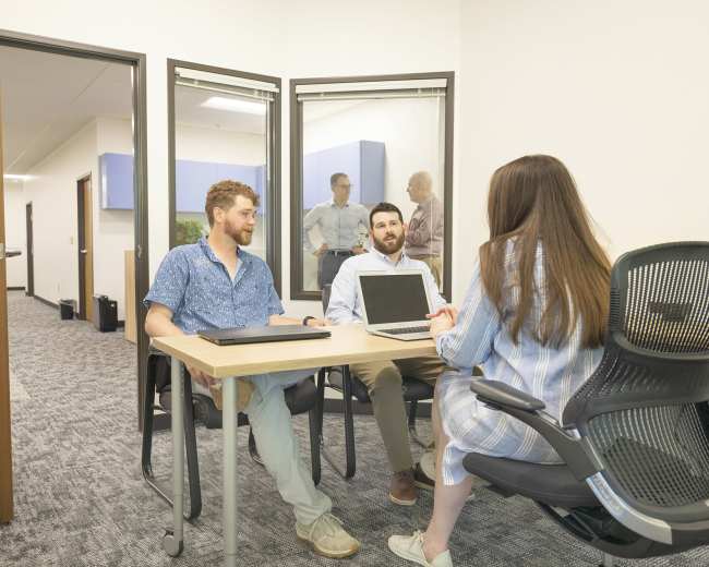 Two men and a woman are seated at a table in an office setting, engaged in a discussion with a laptop open on the table.