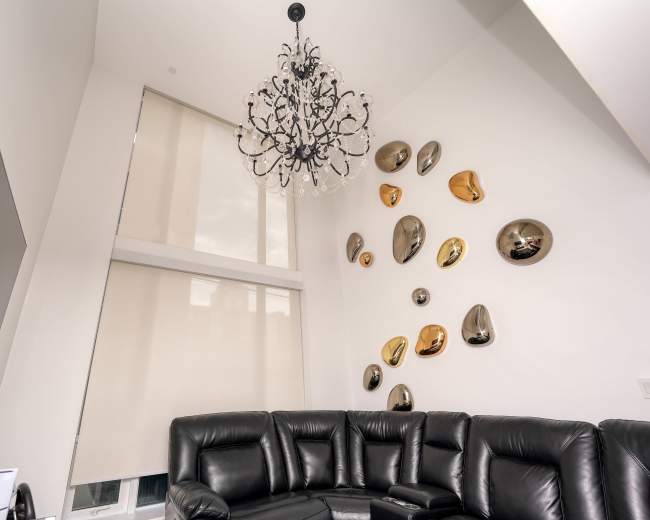 The image shows a modern living room featuring a black leather sectional sofa, a decorative chandelier, and metallic wall art arranged asymmetrically on a white wall.