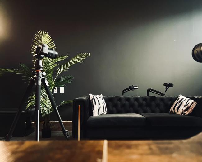 A dark room with a black tufted sofa, two cameras on tripods positioned in front of it, and a potted plant in the background.