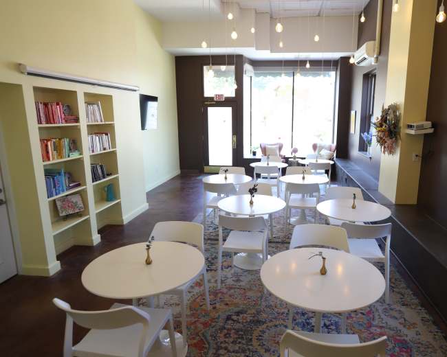 The image shows a brightly lit, sparsely furnished café with white tables, chairs, and a bookshelf, featuring a cozy ambiance created by pendant lights and large windows.