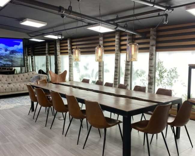 The image shows a modern dining area with a long wooden table surrounded by brown chairs, illuminated by pendant lights, and featuring large windows with a view of greenery.