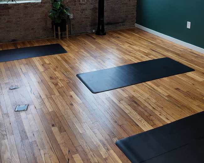 The image shows an open room with wooden floors, large windows, and yoga mats placed on the floor.