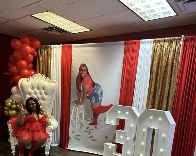 A woman in a red outfit sits on an ornate white chair in a decorated room featuring a large "30" display and festive balloons.