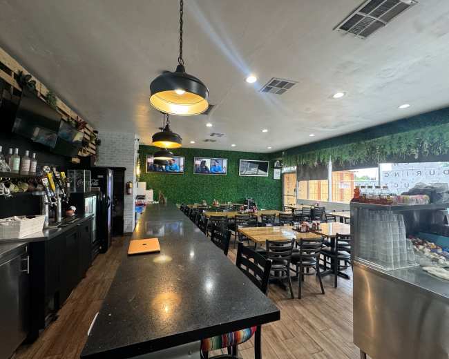 The image shows the interior of a restaurant featuring dark countertops, wooden tables, and green wall decor with several televisions mounted on the walls.