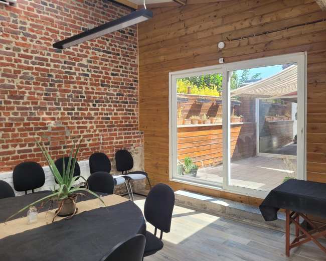 The image shows a bright interior space featuring a wooden wall, a large window overlooking a patio, and a round table surrounded by black chairs.