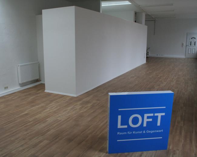 The image shows an empty room with wooden flooring, a blank white wall, and a sign that reads "LOFT Raum für Kunst & Gegenwart."