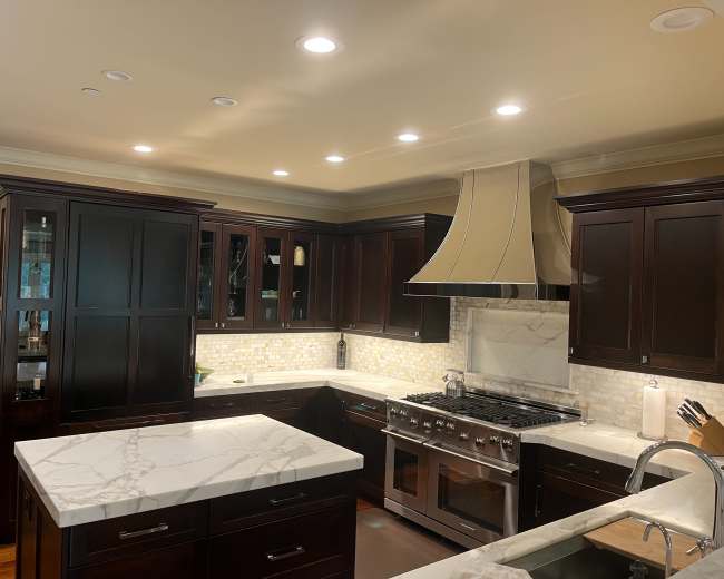 The image shows a modern kitchen featuring dark wooden cabinetry, a large marble countertop, and a stainless steel range hood above a gas stove.