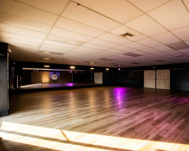 The image shows a spacious dance studio with wooden floors, surrounded by mirrored walls and illuminated by purple lighting.
