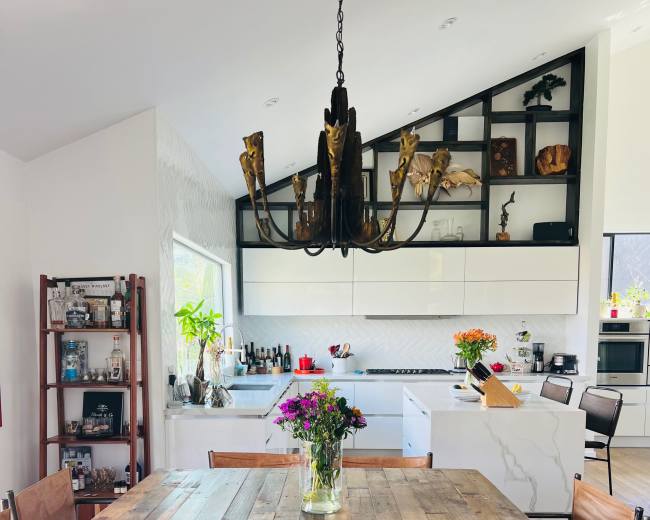 The image shows a modern kitchen and dining area featuring a wooden dining table surrounded by metal and leather chairs, with a chandelier overhead and various kitchen appliances and décor displayed in the background.