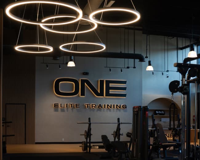 The image shows a modern gym interior featuring exercise equipment and a prominent "ONE ELITE TRAINING" sign on the wall, illuminated by circular light fixtures.