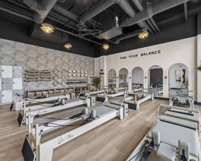 The image shows a modern fitness studio equipped with reformer Pilates machines, large windows, and minimalist decor.