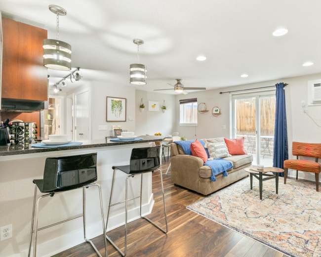 The image shows a modern living space featuring a sofa, dining area, and kitchen bar with stools, illuminated by recessed lighting.
