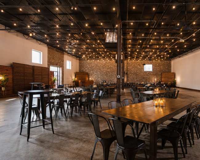 The image shows a spacious event venue with wooden tables and metal chairs, illuminated by overhead string lights, featuring brick walls and a concrete floor.