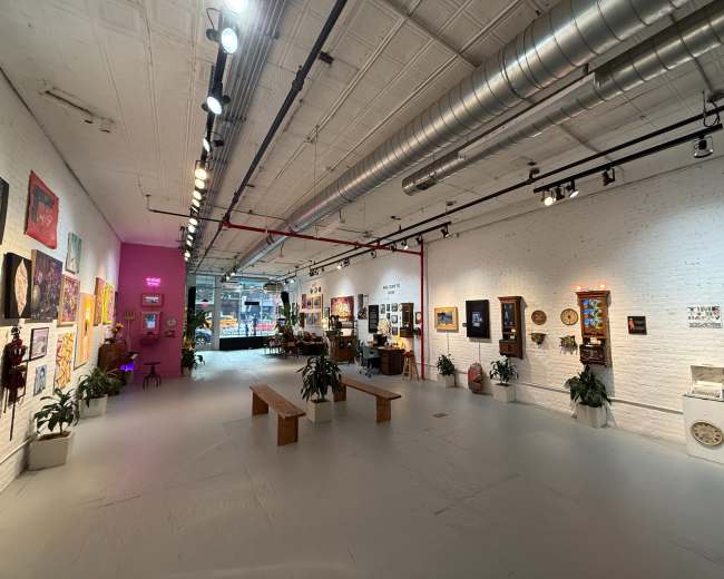 The image shows a spacious art gallery featuring a variety of framed artworks on white brick walls, with plants and wooden benches scattered throughout the room.