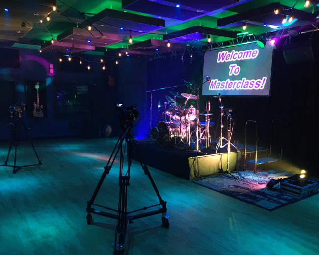 A stage set up for a masterclass, featuring a drum kit, cameras on stands, and a brightly lit sign that reads "Welcome To Masterclass."