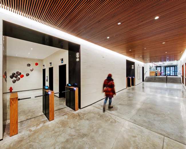 The image shows a modern lobby area with elevators on one side, a concrete floor, and wooden architectural details, while a person walks through the space.