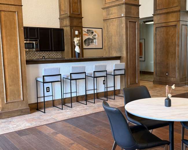 The image shows a modern communal space with a kitchenette featuring bar stools, a round dining table, and wooden pillars.